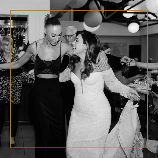 Bride in a long-sleeved gown dances joyfully with a friend in a black dress at the wedding reception.