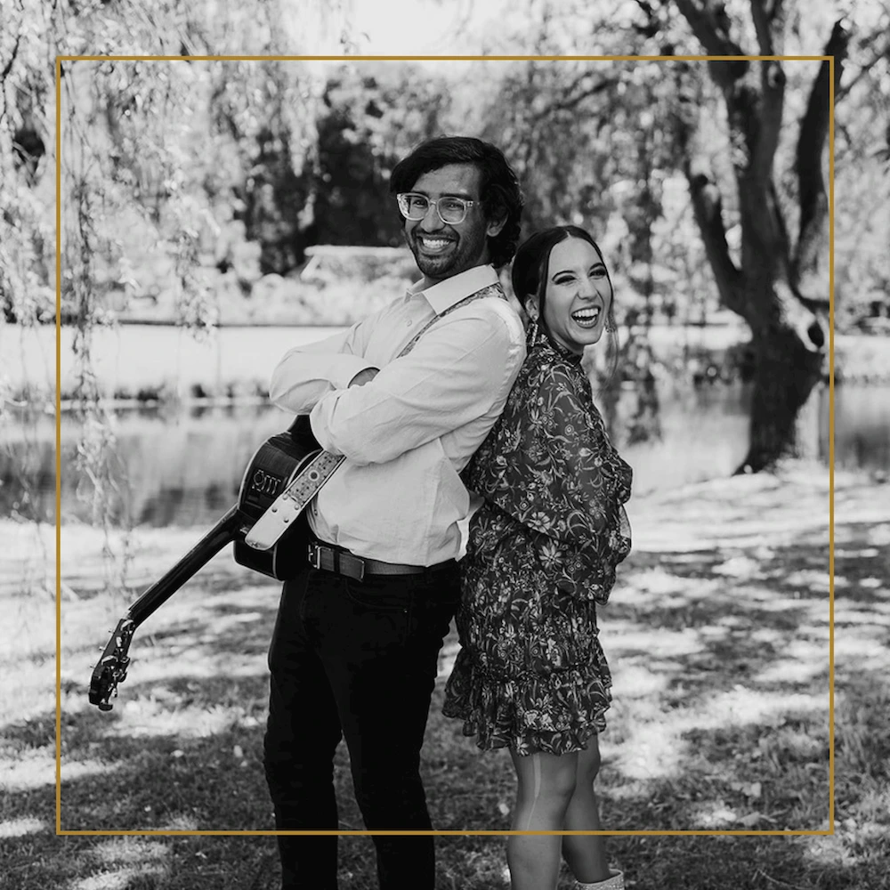 Smiling man with guitar stands back-to-back with woman outdoors by a lake and trees.