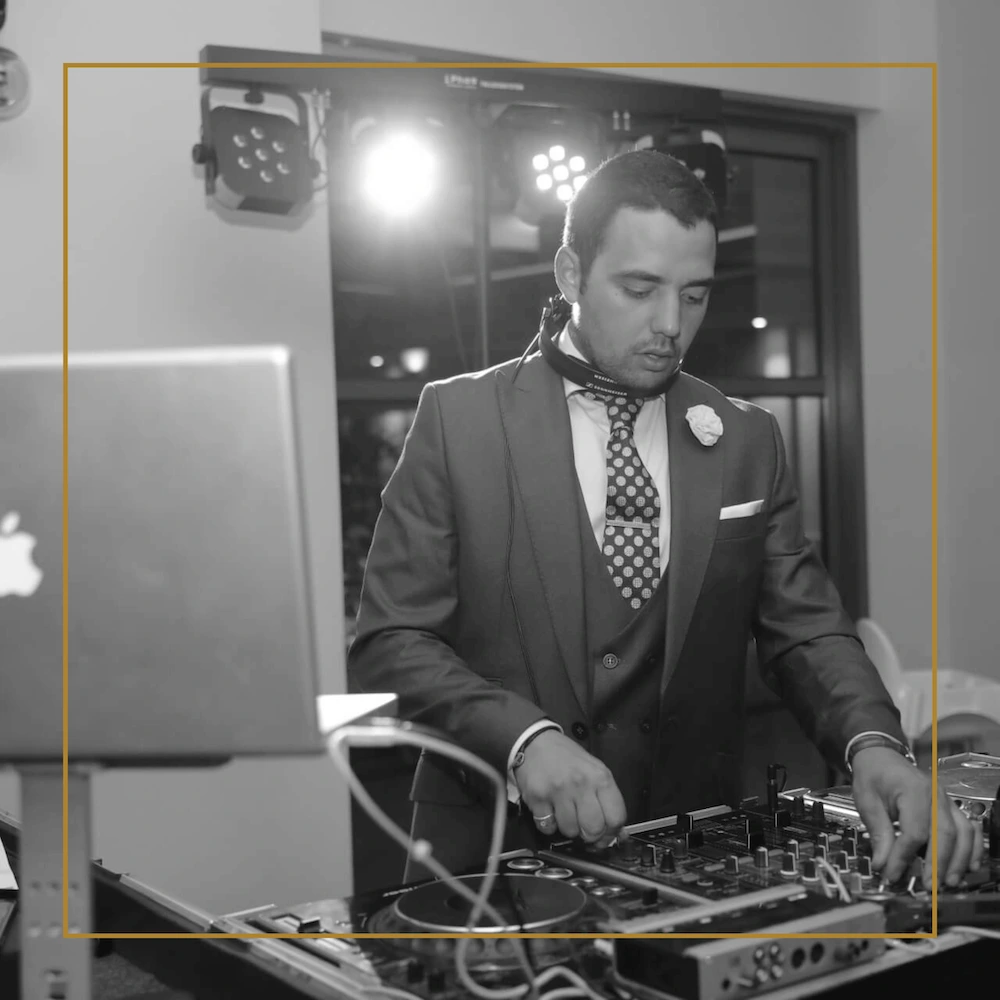 DJ in a suit focused at the decks, wearing headphones around his neck during an event.