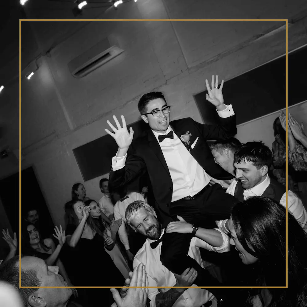 Groom in a tuxedo lifted on friends’ shoulders, smiling and raising hands during a lively wedding reception.