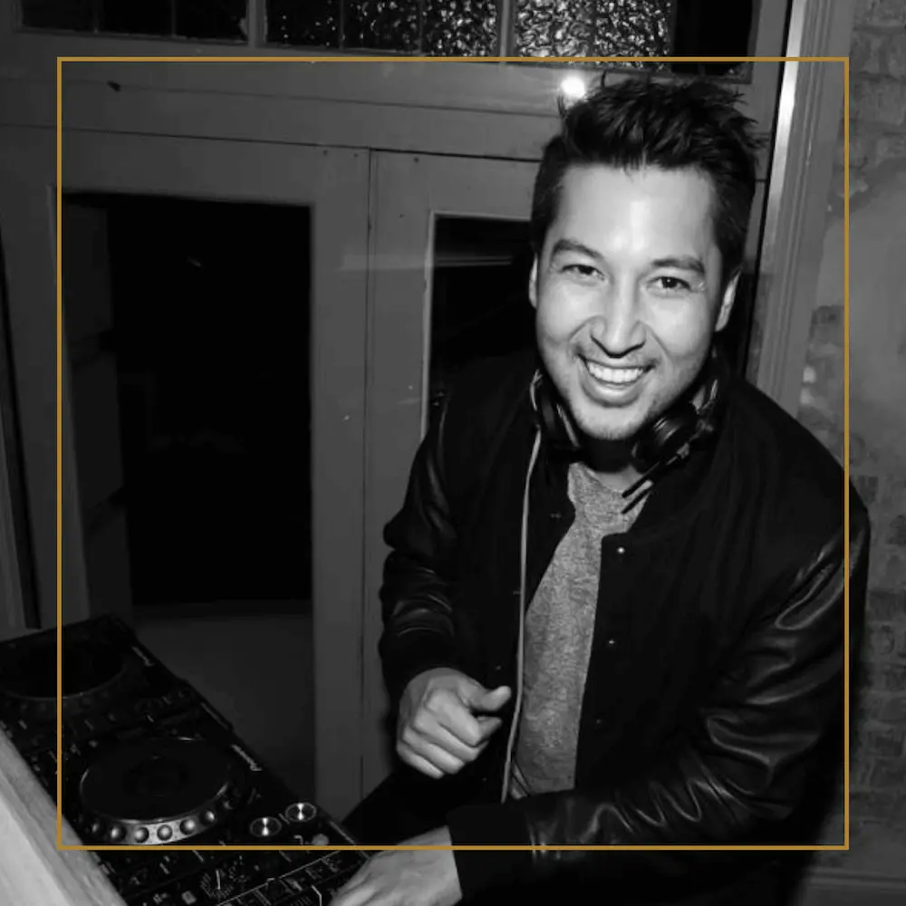 DJ Justin smiling at the decks with headphones around his neck during a set.